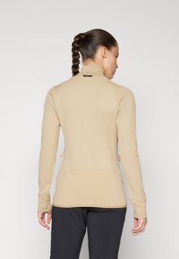 Beige langermet treningstopp med teksturert vaffelmønster, med høy krage og rosa snor detaljer på sidene.