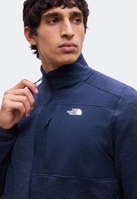 Homme aux cheveux foncés portant une veste zippée North Face bleu marine, tirant la fermeture éclair près du col.