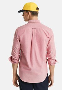Rotkariertes Hemd mit abgerundetem Saum und Knopfdetails. Das Model trägt eine gelbe Kappe, die aus glattem Stoff besteht und hochgekrempelte Ärmel hat.