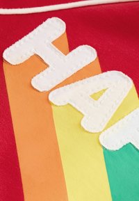 Piros anyag, fehér cérnával hímzett "HAY" felirattal; csíkos háttér narancssárga, sárga és zöld színekben; textúrált, rétegezett design.