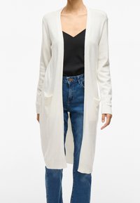 Kvinna iklädd en lång vit öppen cardigan över en svart V-ringad topp och blå högmidjade jeans, stående mot en vit bakgrund.