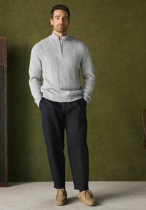 Man stands with hands in pockets wearing light gray cable knit sweater, dark trousers, and tan suede shoes against green wall.