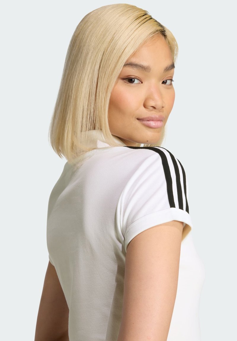 Camisa polo blanca con acentos negros de tres rayas en los hombros. Mangas cortas, textura suave y un diseño ajustado. Modelo mostrado de espaldas.