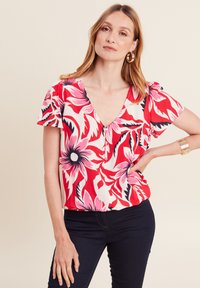 Rote Blumenbluse mit tiefem V-Ausschnitt, kurzen, schwingenden Ärmeln und Knopfdetails. Sie zeigt rosa, schwarze und weiße Blumenmuster auf einem cremfarbenen Hintergrund.