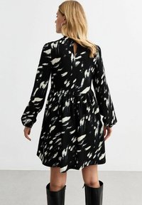 Robe noire avec des motifs abstraits blancs, manches longues, taille froncée, détail en forme de trou de serrure à l'arrière et tissu texturé. Associée à des bottes hautes jusqu'aux genoux.