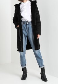 Abrigo largo negro con forro de piel sintética, camisa blanca de botones, jeans claros hasta los tobillos, y botas de tacón negras.
