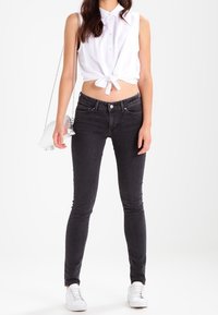 Jeans skinny negros combinados con una camiseta blanca sin mangas anudada a la cintura y un pequeño bolso de bandolera plateado. Zapatillas blancas completan el atuendo.
