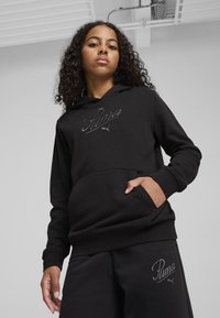 Haut à capuche noir avec poche avant et logo "Puma" brodé. Le tissu semble doux et texturé. Porté avec un pantalon de survêtement noir assorti arborant le logo.