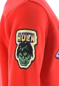 Red sweatshirt sleeve with a sewn patch of The Incredible Hulk's angry green face and bold yellow text above it.
