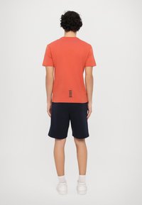 Oransje bomull T-skjorte med rund hals, korte ermer og et særegent horisontalt stripe-detalj nederst på ryggen. Kombinert med marineblå shorts.