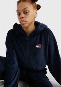 Marineblauwe velours trainingspak met een ritskap. Voorzien van een klein logo-embleem op de borst en een zachte, getextureerde stof.