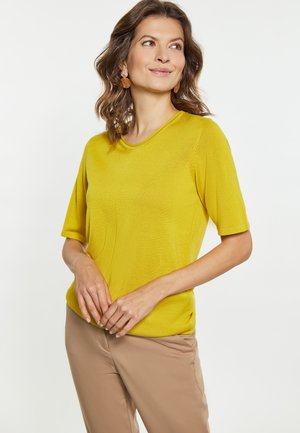 Gul striktrøje med korte ærmer, rund halsudskæring og sideslids, parret med beige bukser. Modellen er accessoriseret med runde øreringe.