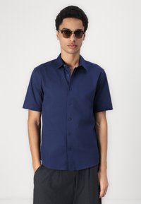 Camisa de botones de manga corta en color navy, hecha de una tela suave. Cuenta con un cuello, botones en la parte delantera y se combina con pantalones oscuros de corte holgado.