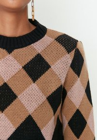 Gros plan sur une personne portant un pull en maille à losanges marron, noir et rose, ainsi qu'une boucle d'oreille pendante en chaîne dorée, sur fond blanc.