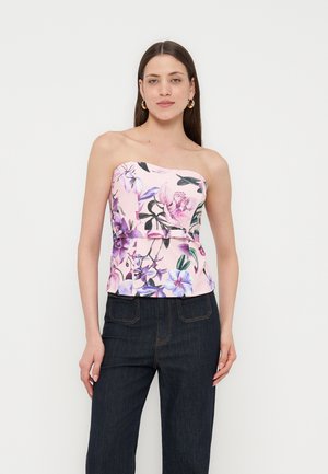 Γυναίκα που φοράει ροζ λουλουδάτο τοπ χωρίς τιράντες συνδυασμένο με σκούρο μπλε τζιν, στέκεται μπροστά σε απλό ανοιχτό φόντο.