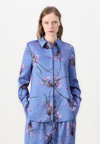 Μπλε μεταξωτό πουκάμισο με λουλουδάτο σχέδιο, με κοφτό γιακά, μακριά μανίκια και δύο πλαϊνές τσέπες. Λείο ύφασμα και λεπτομερές μοτίβο.