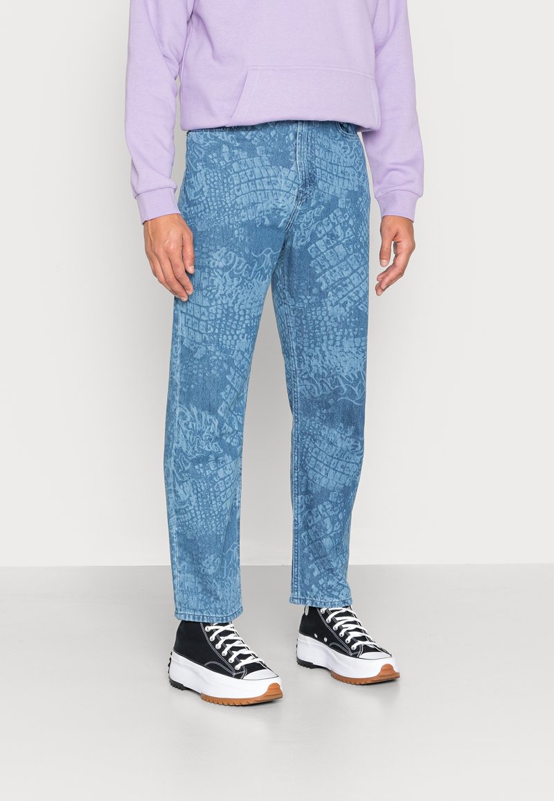 Světlé modré denimové džíny s texturovaným vzorem, rovný střih. Vpárovány s fialovou mikinou a černými teniskami s bílými detaily.