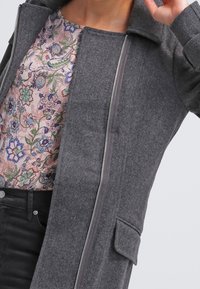 Grijze wollen jas met een rits aan de voorkant, kraag en zijzakken. Draagt over een bloemdessin top in roze en blauwe tinten.