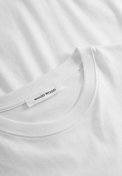 Vit bomull t-shirt med rund hals, med en tagg märkt "WOOD WOOD." Tyget verkar jämnt med en subtil textur.