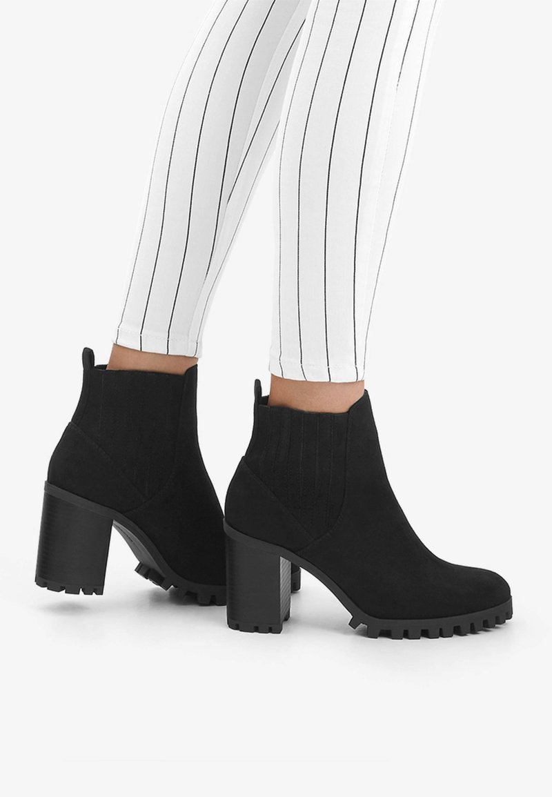 Black suede ankle boots with elastic side panels, chunky block heels, and textured rubber soles, paired with white striped leggings.