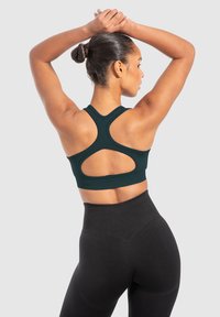 Tummanvihreä urheiluliivi kilpailijaelämällä, jossa on leikkauksella varustettu muotoilu, yhdistettynä korkeavyötäröisiin mustiin leggingseihin, jotka ovat sileäpintaiset.