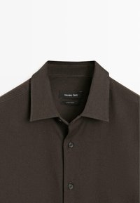 Brown, short-sleeve button-up shirt with a collar, featuring a textured fabric and a logo label inside the neckline. Four black buttons visible.