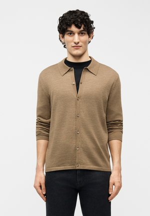 Jeune homme aux cheveux bouclés portant un cardigan marron boutonné sur une chemise noire, debout devant un fond clair uni.