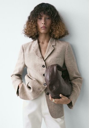 Persona con capelli ricci fino alle spalle, che indossa un blazer beige e pantaloni bianchi, mentre tiene una grande borsa piegata in pelle marrone.