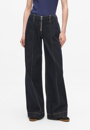 Dark blue denim wide-leg pants with front zipper, featuring contrasting yellow stitching and deep side pockets. Smooth texture, high waist design.