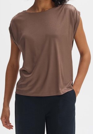 Kobieta nosząca luźną brązową bluzkę z krótkim rękawem, wsuniętą na zewnątrz na czarne, fakturowane spodnie, stoi z jedną ręką w kieszeni na białym tle.