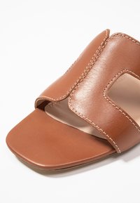 Brown leather sandal featuring a wide strap with cutouts, a smooth texture, and fine stitching. Square toe design with a minimalistic shape.