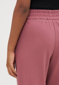 Růžové elastické kalhoty s hladkou texturou, které mají volný střih. Záběr zahrnuje část ruky opřenou o nohu.