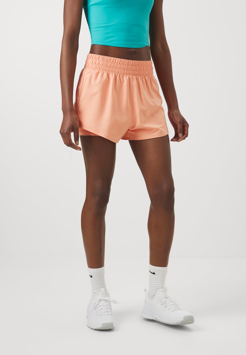 Shorts pêches avec une taille élastique, présentant une coupe décontractée et une texture lisse, associés à des baskets blanches et des chaussettes de sport.
