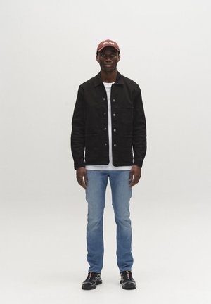 Mand iført mørkerød kasket, sort knappet jakke, hvid skjorte, lyseblå jeans og sorte sneakers, stående mod enkel hvid baggrund.