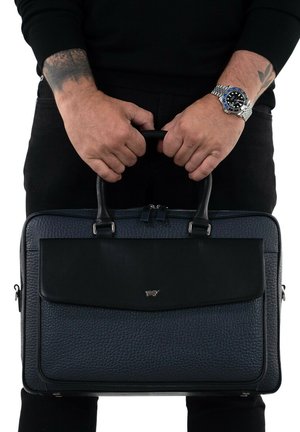 Schwarze Lederaktentasche mit einer strukturierten, genarbten Oberfläche, versehen mit einer eleganten Klappe und einem Metalllogo, gehalten von zwei Händen, an denen eine Uhr sichtbar ist.