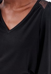 Blouse noire à col en V avec tissu doux, encolure froncée et accents en maille transparente sur les épaules pour un contraste texturé.