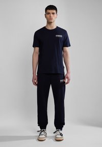 Navy Baumwoll-T-Shirt und passende Jogginghose, mit einem Logo-Print auf der Brust und der Tasche. Die Schuhe sind weiß mit dunklen Akzenten und Schnürsenkeln.