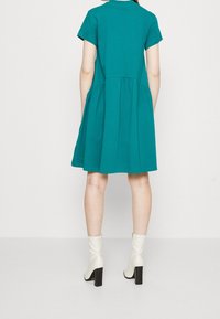 Tyrkysové šaty s krátkým rukávem, s přiléhavým korzetem a rozšířenou sklady. Mají kulatý výstřih a jsou vyrobeny z měkké bavlny.