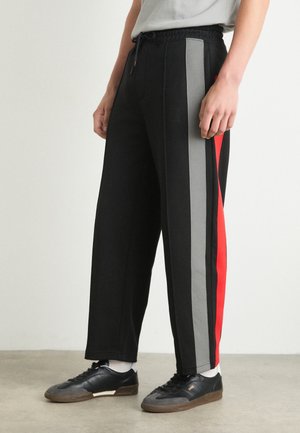 Black sweatpants with gray and red side stripes, featuring a drawstring waistband and a relaxed fit, paired with black sneakers.