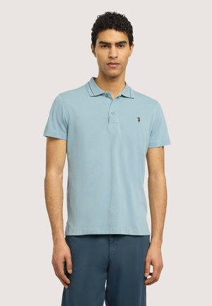 Młody mężczyzna z krótkimi kręconymi włosami, ubrany w jasnoniebieską koszulkę polo i ciemnoniebieskie spodnie, stoi na jednolitym jasnym tle.