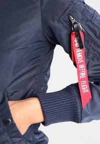 Sötétkék bomber dzseki bordázott mandzsettákkal és "Remove Before Flight" feliratos címkével. Jellemzője a karon található cipzáras zseb és a sima, fényes anyag.