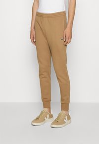 Pantaloni de trening maro, dintr-un material moale, având o talie elastică, picioare conice și un mic logo verde. Asortat cu adidași bej.