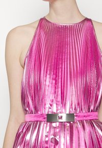 Femme portant une robe rose brillante, plissée, sans manches, avec une ceinture assortie dotée d'une boucle métallique carrée.