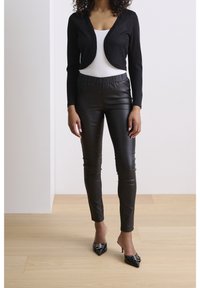 Kobieta ubrana w czarny krótkookresowy kardigan, biały top, czarne skórzane spodnie i czarne szpilki z ostro zakończonym noskiem, stojąca na jasnej drewnianej podłodze.