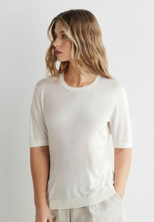 Kvinde med bølget blondt hår iført en enkel hvid kortærmet striktrøje, stående med den ene hånd i lommen, kigger mod venstre mod en hvid baggrund.