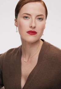 Złota naszyjnik z pięcioma okrągłymi perłami, noszony na brązowym swetrze z dekoltem w serek. Do kompletu odpowiadają kolczyki z perłami wiszącymi. Gładkie tekstury.