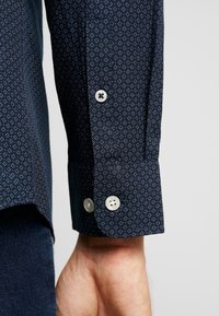 Marinfärgad skjorta med lång ärm och små vita diamantsymboler, vita knappar, och synlig manschett nära en persons handled.