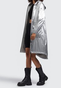 Srebrny, wodoodporny długi płaszcz z kapturem, zapinany na zamek, z regulowanym sznurkiem. Nosi się go na czarny crop top i dopasowaną spódnicę, do tego czarne bojówki.