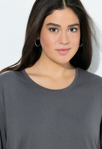 Grijze t-shirt met korte mouwen en een ronde halslijn, zachte textuur en opvallende stiksels. Model met lang donker haar en oorringen.