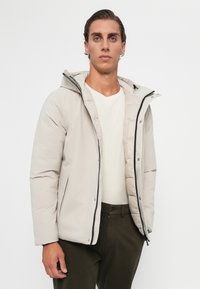 Giacca trapuntata color beige chiaro con cappuccio, zip frontale e bottoni a pressione. Tessuto liscio, con zip nera e dettagli di cucitura interna.